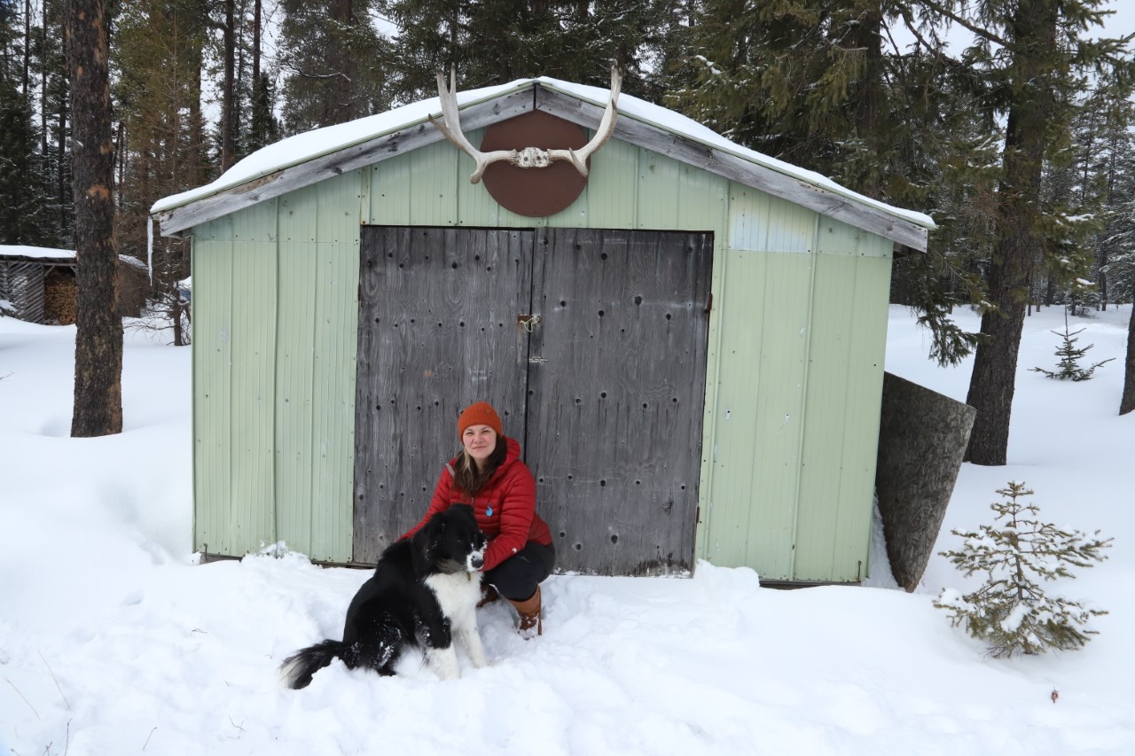 Valérie et son compagnon canin devant un cabanon orné d'un panache d'orignal prennent la pause. Il y a de la neige au sol et une forêt les entoure. 